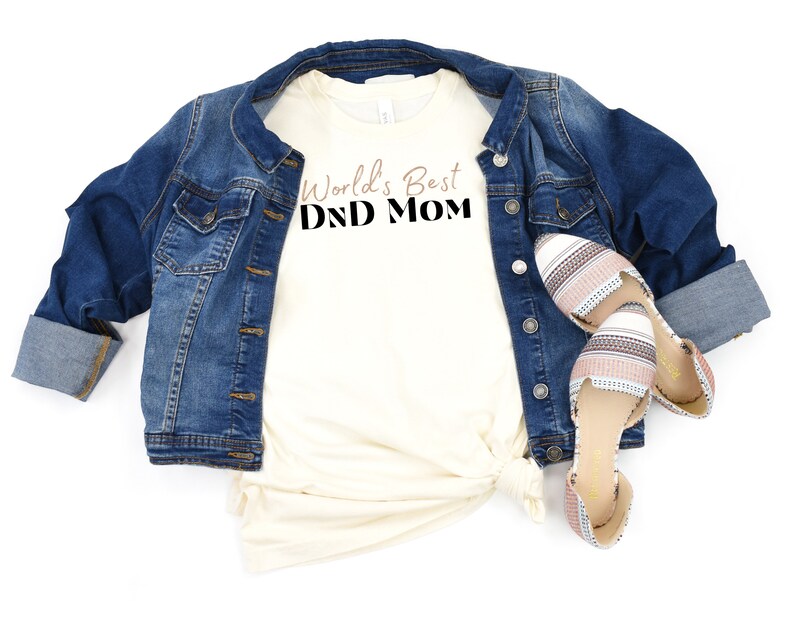 May include: A white t-shirt with the text "World's Best DND Mom" in black and brown lettering. The shirt is paired with a denim jacket and striped flats.