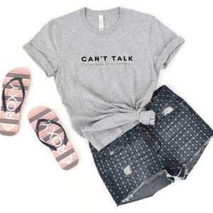 May include: A gray t-shirt with the text "CAN'T TALK I'm working on my backstory" printed on the front. The shirt is paired with denim shorts and pink flip-flops.