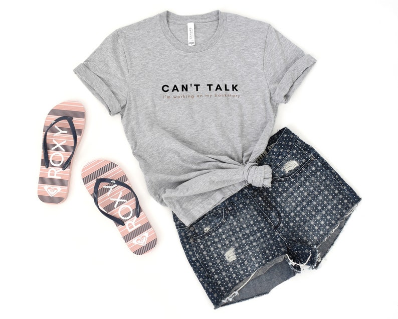 May include: A gray t-shirt with the text "CAN'T TALK I'm working on my backstory" printed on the front. The shirt is paired with denim shorts and pink flip-flops.