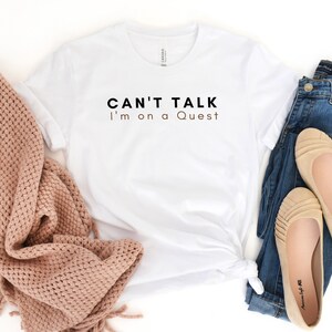 May include: White t-shirt with the text "CAN'T TALK I'm on a Quest" in black and brown lettering.