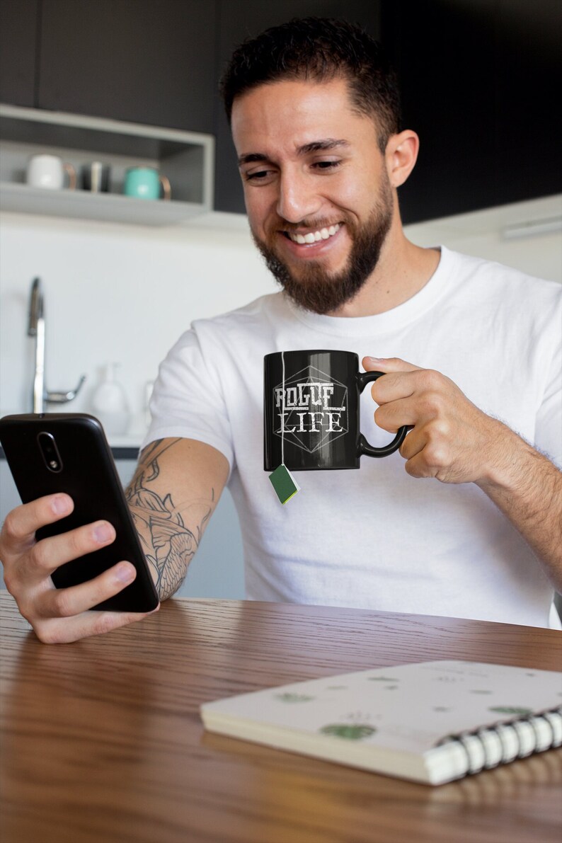 Op de afbeelding: Een persoon houdt een zwarte mok vast met de tekst "Rogue Life" erop gedrukt. De mok is gevormd als een 20-zijdige dobbelsteen. De persoon glimlacht en kijkt naar een smartphone.