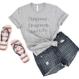 May include: A gray t-shirt with the text "Dungeons Dragons Mom Life" and a small graphic of a dice. The shirt is knotted and paired with denim shorts and pink flip-flops.