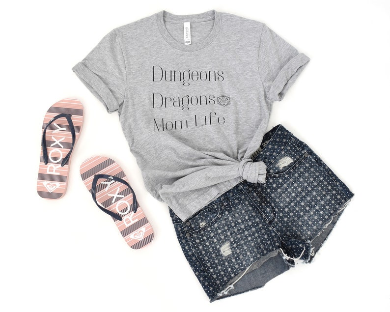 May include: A gray t-shirt with the text "Dungeons Dragons Mom Life" and a small graphic of a dice. The shirt is knotted and paired with denim shorts and pink flip-flops.