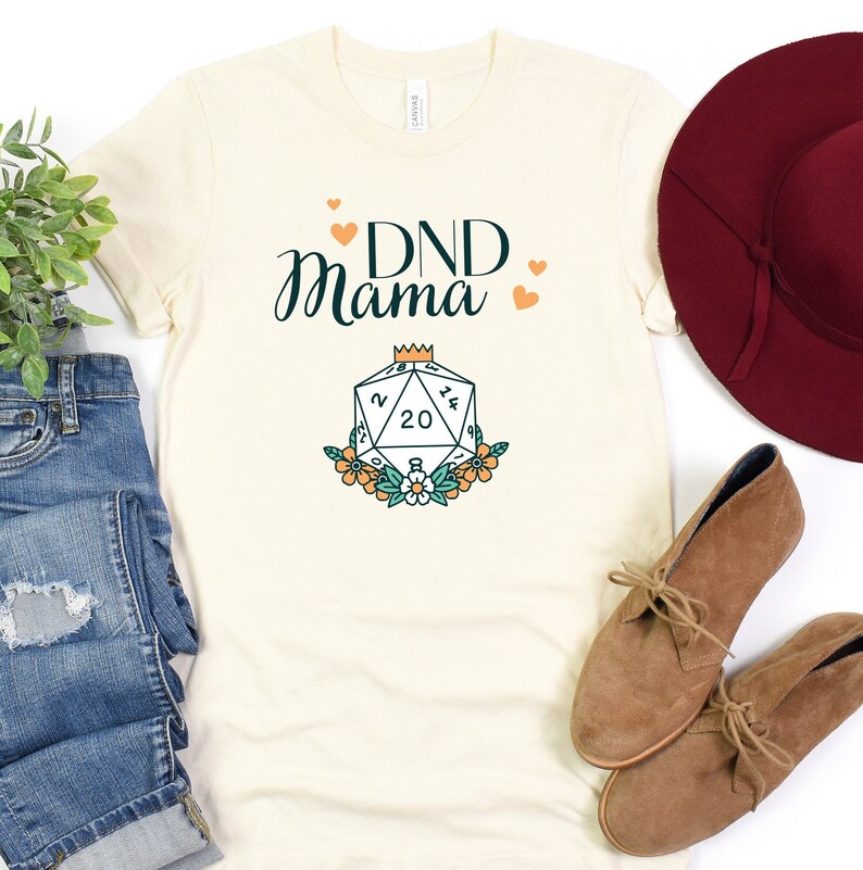 May include: A white t-shirt with the text "DND Mama" in green and black lettering. A stylized illustration of a twenty-sided die with a crown on top is below the text. The die is surrounded by orange and white flowers.