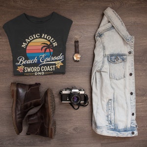Pode incluir: Uma camiseta preta com um gr&aacute;fico de p&ocirc;r do sol retr&ocirc; e o texto "Magic Hour Beach Episode Sword Coast Fantasy DND Beach". A camiseta est&aacute; colocada sobre uma superf&iacute;cie de madeira com um par de botas de couro marrom, uma c&acirc;mera, um rel&oacute;gio e uma jaqueta jeans.