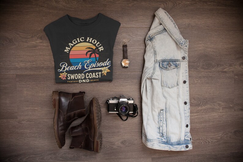 Pode incluir: Uma camiseta preta com um gr&aacute;fico de p&ocirc;r do sol retr&ocirc; e o texto "Magic Hour Beach Episode Sword Coast Fantasy DND Beach". A camiseta est&aacute; colocada sobre uma superf&iacute;cie de madeira com um par de botas de couro marrom, uma c&acirc;mera, um rel&oacute;gio e uma jaqueta jeans.