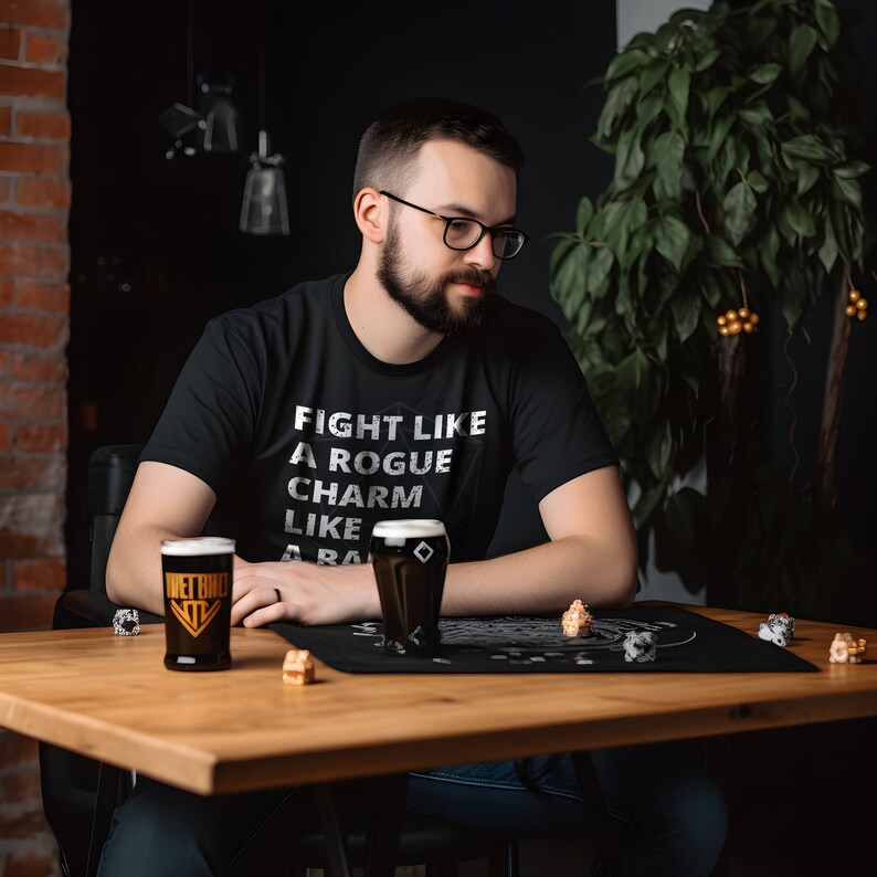 May include: A person wearing a black t-shirt with the text "Fight like a rogue, charm like a bard" sits at a table with two glasses of dark beer. The table has a black gaming mat with dice and other game pieces.