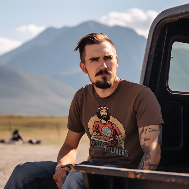 May include: A man wearing a brown t-shirt with a graphic of a person holding a pizza and the text "Jesus Delivers". He has a beard and tattoos on his arm.