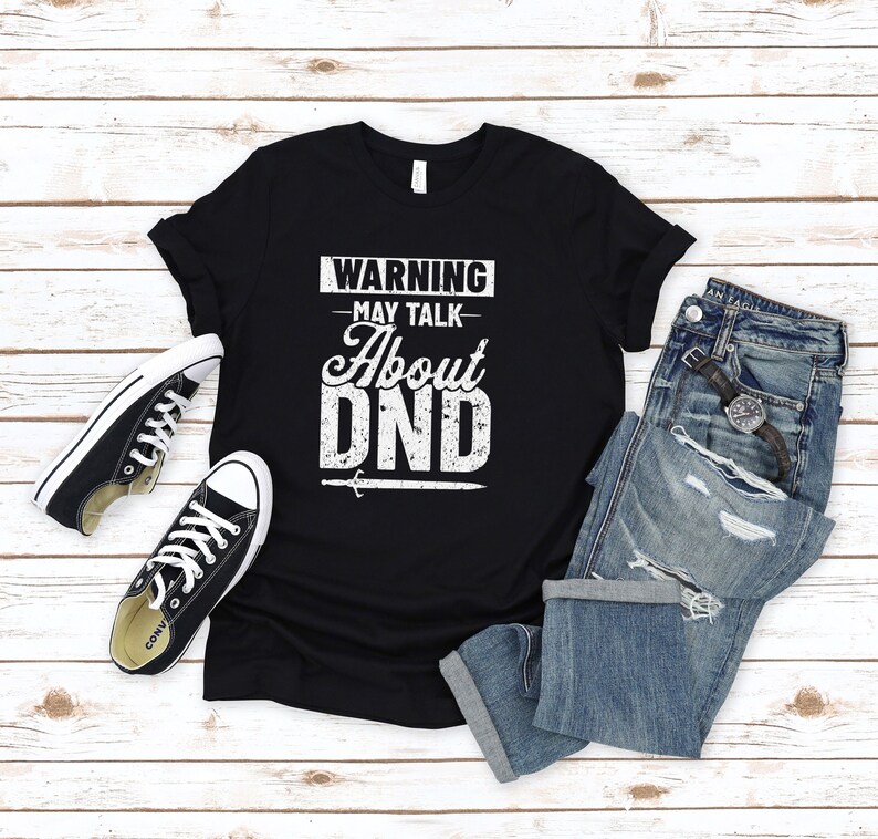 May include: Black t-shirt with white text that reads "WARNING - MAY TALK ABOUT DND" with a sword graphic. The shirt is on a white wood background with a pair of black sneakers and blue jeans.