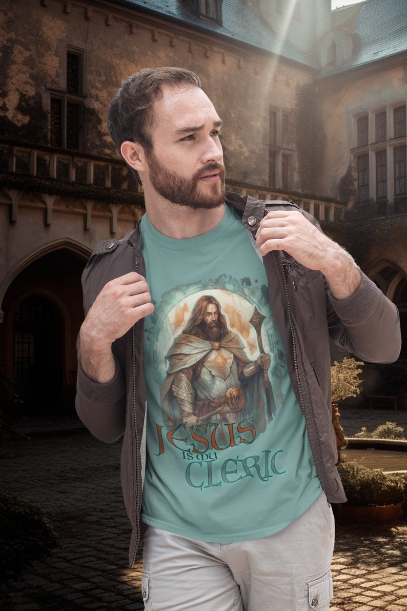May include: A light blue t-shirt with a graphic of a man in armor holding a staff. The text "Jesus is my Cleric" is printed on the shirt.