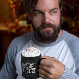 Op de afbeelding: Een zwarte keramische mok met een wit ontwerp met de tekst "Rogue Life" en een witte geometrische vorm. De mok is gevuld met een romige drank die met kaneel is bestrooid.