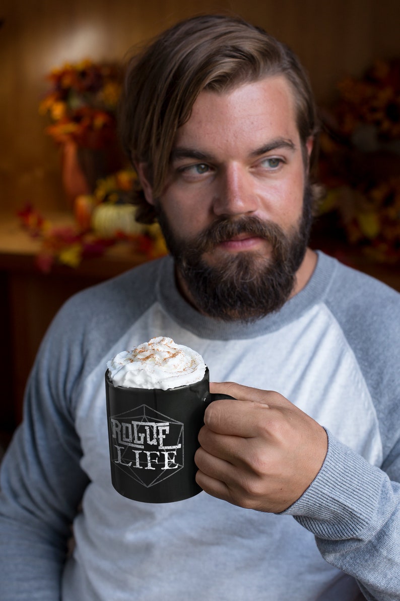 Op de afbeelding: Een zwarte keramische mok met een wit ontwerp met de tekst "Rogue Life" en een witte geometrische vorm. De mok is gevuld met een romige drank die met kaneel is bestrooid.