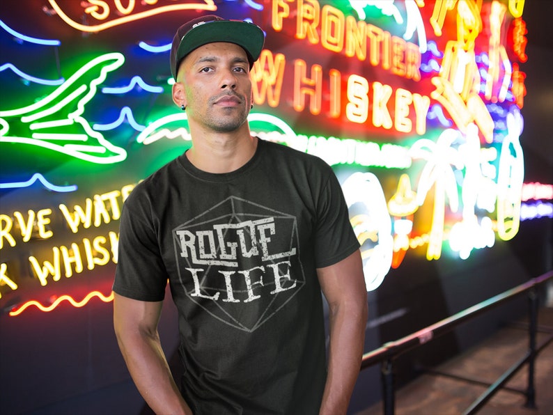 May include: A black t-shirt with a white graphic of a 20-sided die with the text "Rogue Life" inside. The person wearing the shirt is standing in front of a neon sign that says "Frontier Whiskey".