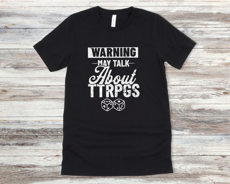 May include: Black t-shirt with white text that reads "WARNING - MAY TALK ABOUT TTRPGS" and two white dice.