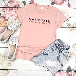 May include: A pink t-shirt with the text "Can't Talk I'm on a Quest" printed in black.