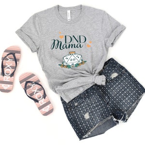 May include: Gray t-shirt with the text "DND Mama" and a green 20-sided die with a crown on top. The die is surrounded by flowers.