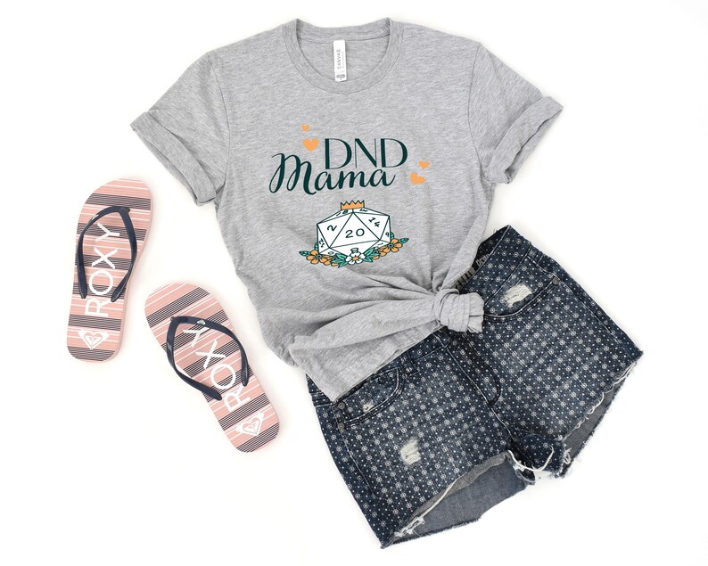 May include: Gray t-shirt with the text "DND Mama" and a green 20-sided die with a crown on top. The die is surrounded by flowers.