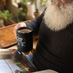 Op de afbeelding: Een man met een lange witte baard houdt een zwarte koffiemok vast met de tekst "League of Coffee" erop gedrukt. Hij zit en leest een tijdschrift.