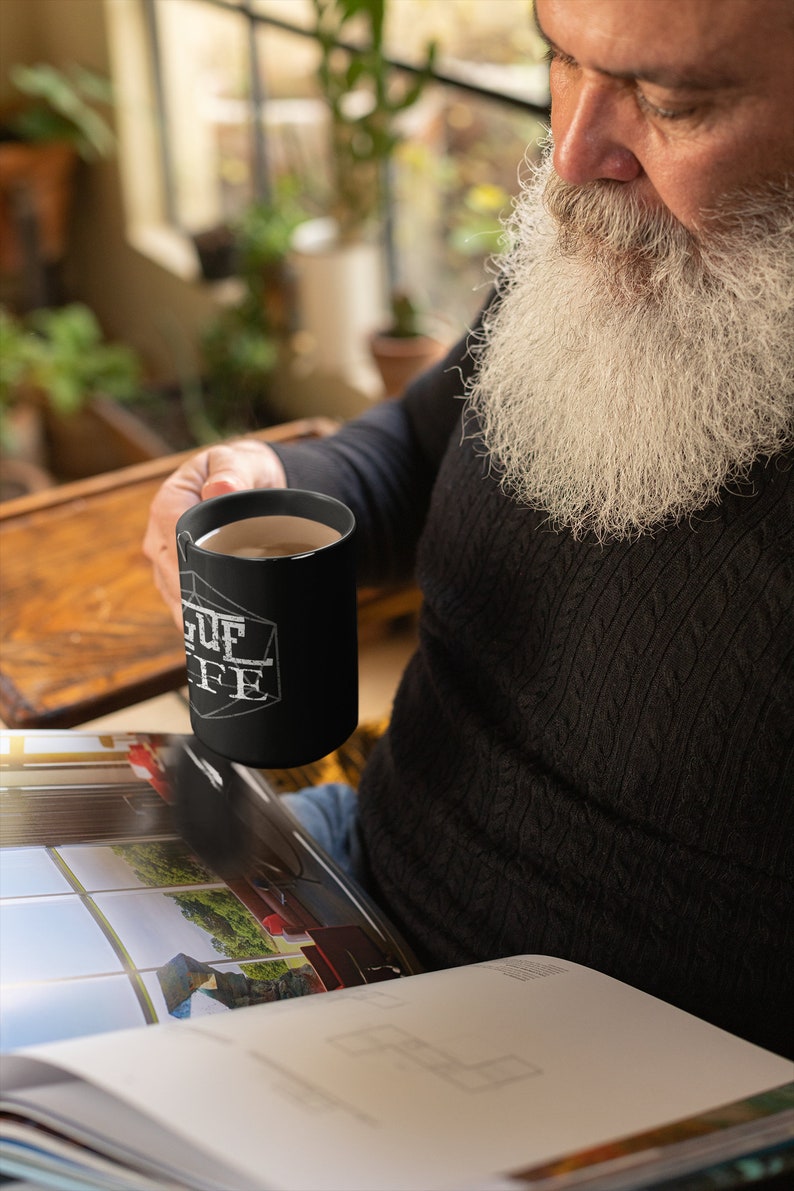 Op de afbeelding: Een man met een lange witte baard houdt een zwarte koffiemok vast met de tekst "League of Coffee" erop gedrukt. Hij zit en leest een tijdschrift.