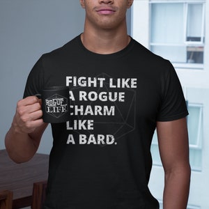 Op de afbeelding: Een zwart T-shirt met witte tekst die "Fight like a rogue, charm like a bard." zegt. Een zwarte mok met witte tekst die "Rogue Life" zegt, wordt vastgehouden door een persoon.