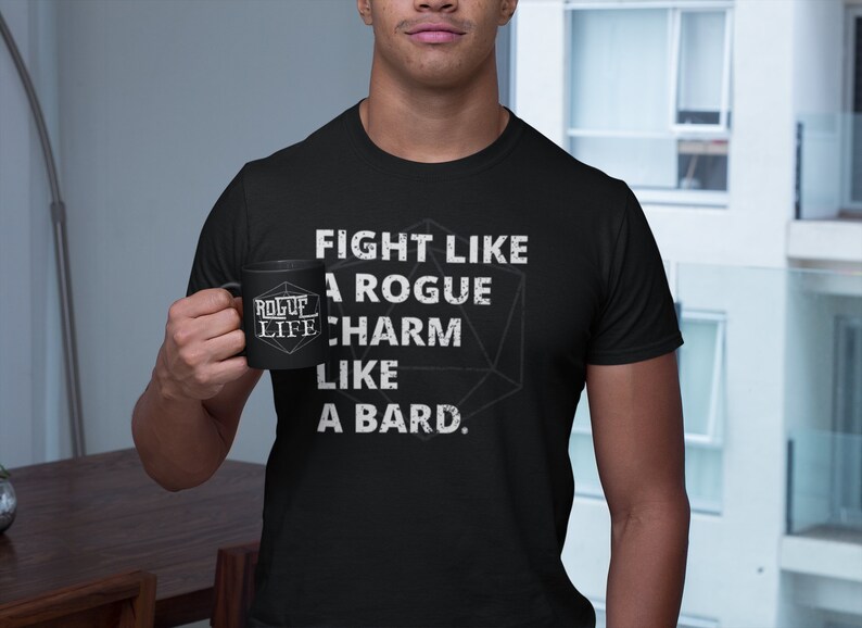 Op de afbeelding: Een zwart T-shirt met witte tekst die "Fight like a rogue, charm like a bard." zegt. Een zwarte mok met witte tekst die "Rogue Life" zegt, wordt vastgehouden door een persoon.