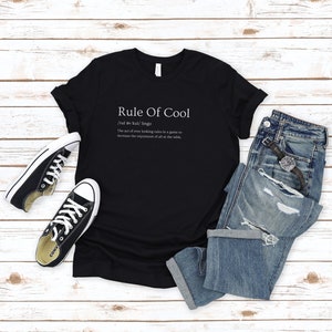 May include: Black t-shirt with white text that reads "Rule Of Cool /rul av kul/ lingo The act of over looking rules in a game to increase the enjoyment of all at the table."