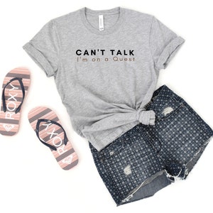 May include: A grey t-shirt with the text "CAN'T TALK I'm on a Quest" in black and brown. The shirt is paired with denim shorts and pink flip-flops with the word "ROXY" printed on them.
