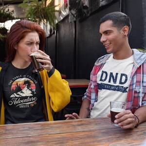 The table is calling adventure awaits dnd t-shirt design on a trendy young woman sitting at a hipster bar drinking craft beer with a dude in a flannel and a "DND EST 1974" t-shirt