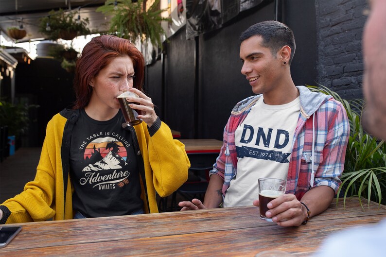 The table is calling adventure awaits dnd t-shirt design on a trendy young woman sitting at a hipster bar drinking craft beer with a dude in a flannel and a "DND EST 1974" t-shirt