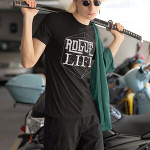 May include: A person wearing a black t-shirt with the text "Rogue Life" printed on it in white. The text is inside a white outline of a 20-sided die. The person is wearing a green jacket and holding a black katana sword over their shoulder.