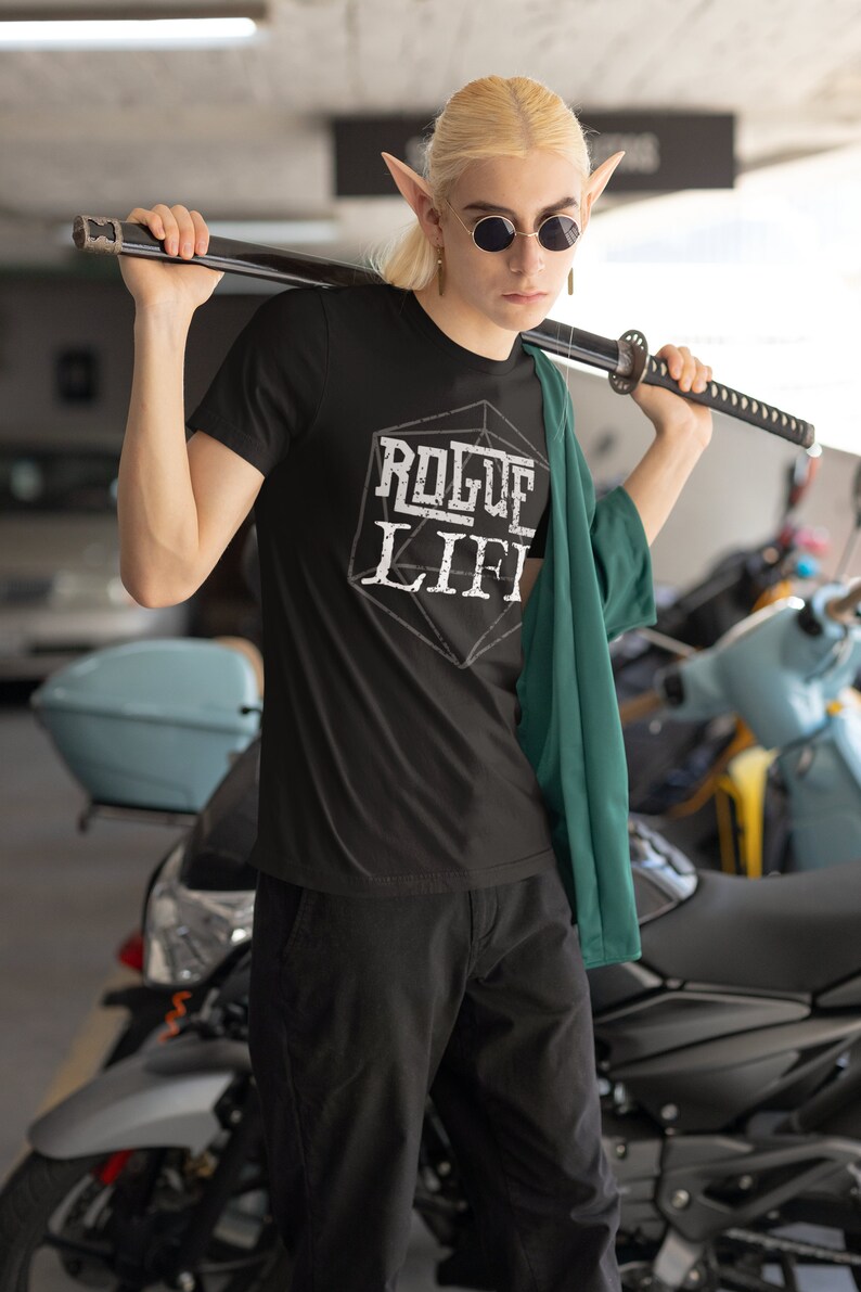 May include: A person wearing a black t-shirt with the text "Rogue Life" printed on it in white. The text is inside a white outline of a 20-sided die. The person is wearing a green jacket and holding a black katana sword over their shoulder.