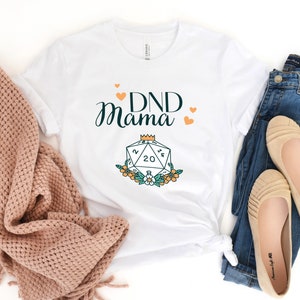 May include: White t-shirt with the text "DND Mama" in black and green lettering. A stylized illustration of a twenty-sided die with a crown on top is below the text.