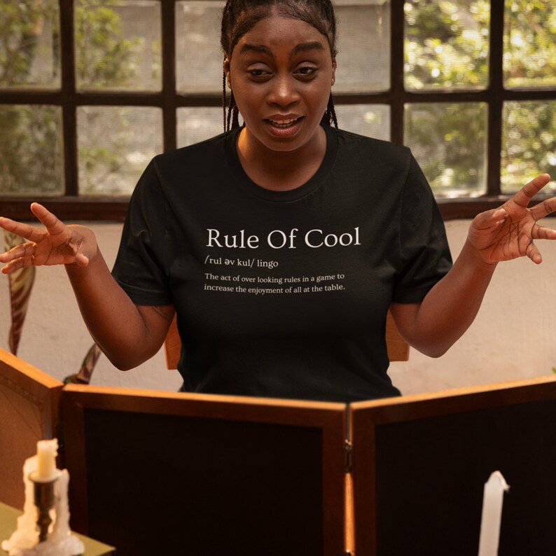 May include: A person wearing a black t-shirt with white text that reads "Rule Of Cool /rul av kul/ lingo The act of over looking rules in a game to increase the enjoyment of all at the table."