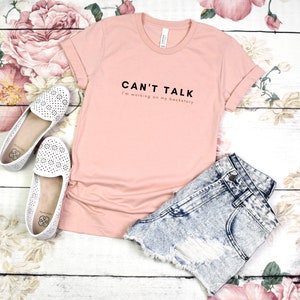 May include: A pink t-shirt with the text "CAN'T TALK I'm working on my backstory" printed on the front. The shirt is laid out on a white wooden surface with floral patterns.