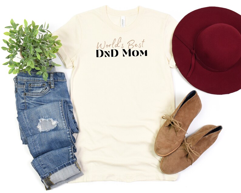 May include: A beige t-shirt with the text "World's Best DND Mom" printed in black on the front. The shirt is laid flat on a white surface with a pair of brown suede boots, a pair of blue jeans, and a burgundy wide-brimmed hat.