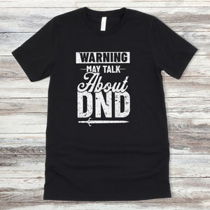 May include: Black t-shirt with white text that reads "WARNING - MAY TALK ABOUT DND" with a sword graphic below the text.