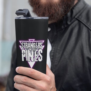 K&ouml;nnte beinhalten: Ein schwarzer Edelstahlbecher mit einer lila und wei&szlig;en Dreiecksgrafik, auf der "Strangers in the Pines" steht.