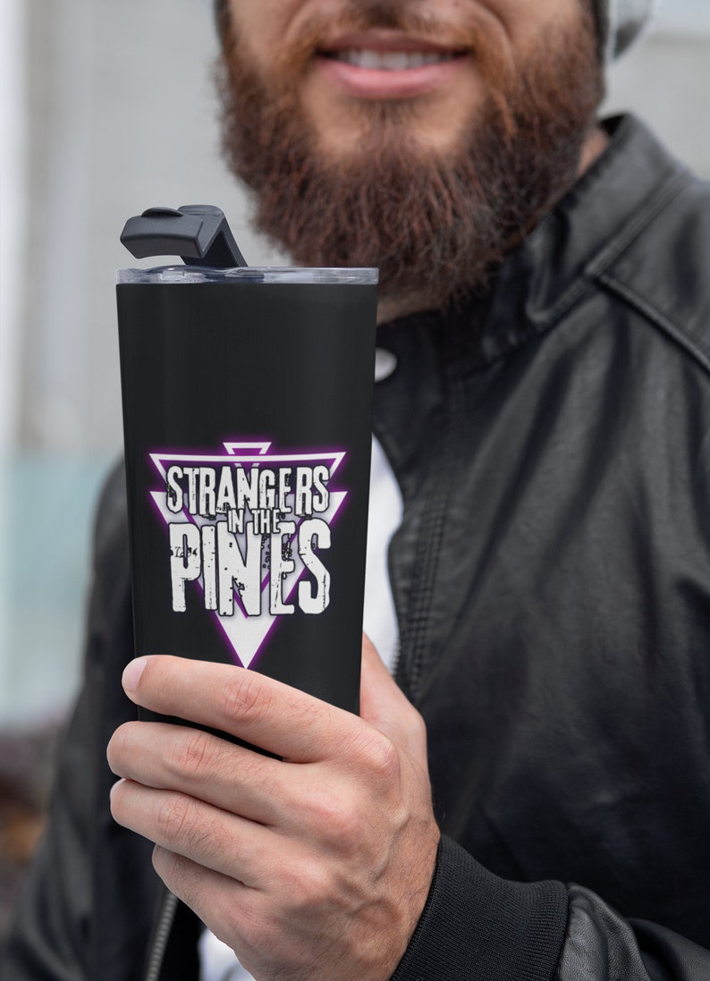 K&ouml;nnte beinhalten: Ein schwarzer Edelstahlbecher mit einer lila und wei&szlig;en Dreiecksgrafik, auf der "Strangers in the Pines" steht.