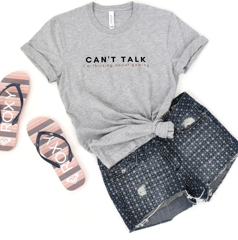 K&ouml;nnte beinhalten: Ein graues T-Shirt mit dem Text "CAN'T TALK I'm thinking about gaming." in schwarzer und ros&eacute;goldener Schrift. Das Shirt wird mit Jeans-Shorts und pinkfarbenen und blauen Flip-Flops kombiniert.
