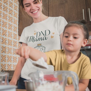 May include: A woman wearing a white t-shirt with the text "DND Mama" and a gold crown above a green dice. She is in a kitchen with a child wearing a yellow shirt. They are both holding spoons and a bowl of white powder.