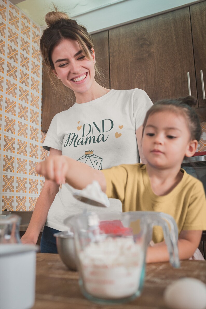 May include: A woman wearing a white t-shirt with the text "DND Mama" and a gold crown above a green dice. She is in a kitchen with a child wearing a yellow shirt. They are both holding spoons and a bowl of white powder.