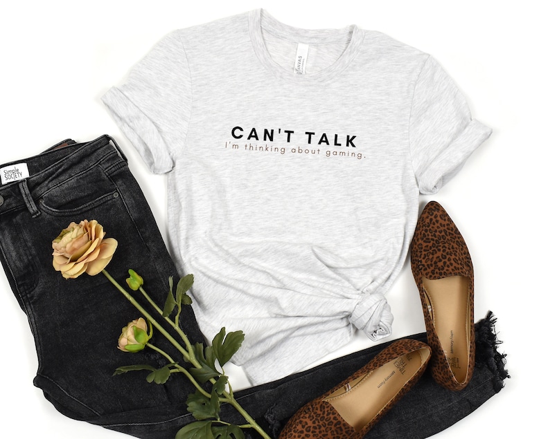 K&ouml;nnte beinhalten: Ein graues T-Shirt mit dem Text "CAN'T TALK I'm thinking about gaming." in Schwarz und Braun auf der Vorderseite. Das Shirt wird mit einer schwarzen Jeans und Leoparden-Flachschuhen kombiniert.