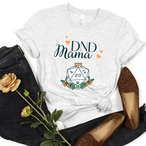 May include: A gray t-shirt with the text "DND Mama" and a stylized illustration of a twenty-sided die with a crown on top and flowers around it.