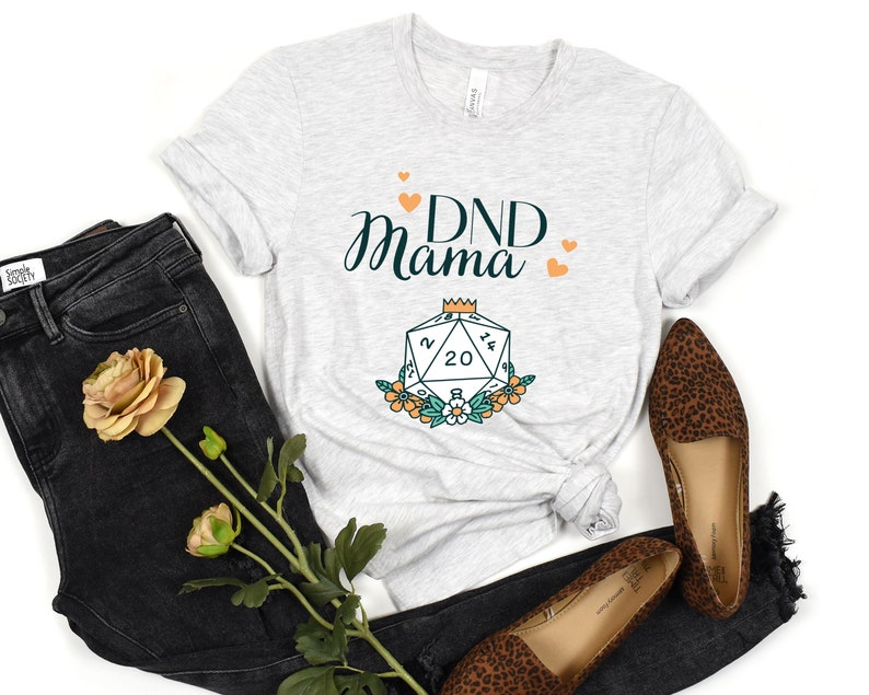 May include: A gray t-shirt with the text "DND Mama" and a stylized illustration of a twenty-sided die with a crown on top and flowers around it.