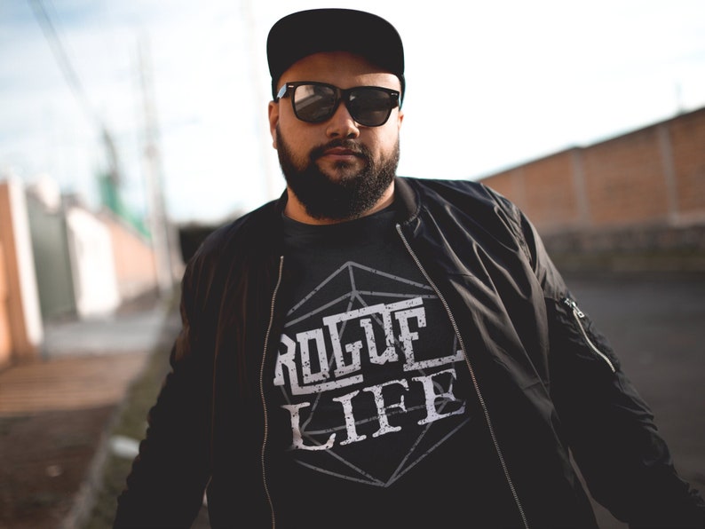 May include: A man wearing a black bomber jacket with a black t-shirt underneath. The t-shirt has a white graphic design of a geometric shape with the words "ROGUE LIFE" inside.