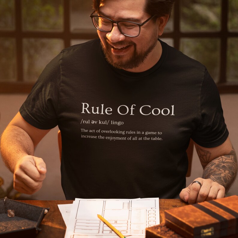 May include: A black t-shirt with white text that reads "Rule Of Cool /rul av kul/ lingo The act of overlooking rules in a game to increase the enjoyment of all at the table."