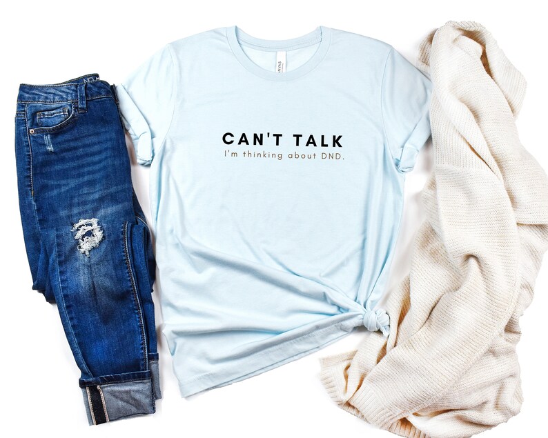 May include: A light blue t-shirt with the text "CAN'T TALK I'm thinking about DND."  The shirt is paired with blue jeans and a white knitted cardigan.