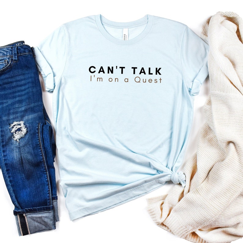 May include: A light blue t-shirt with the text "CAN'T TALK I'm on a Quest" in black and brown lettering.