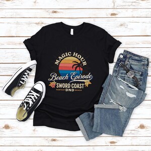 Pode incluir: Camiseta preta com um gr&aacute;fico de p&ocirc;r do sol retr&ocirc; e o texto "Magic Hour Beach Episode Sword Coast Fantasy DND Beach".