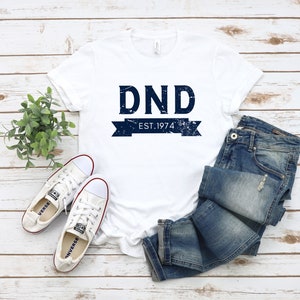 May include: White t-shirt with a blue distressed graphic that says "DND EST. 1974". The shirt is laying on a white wooden surface with a pair of white sneakers and blue jeans.